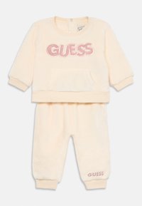 Vauvamallin kreemivärvi plush pitkähihainen collegepaita ja housut, joissa on vaaleanpunainen paljetein koristeltu "GUESS" logo rinnassa ja housunlahkeessa.