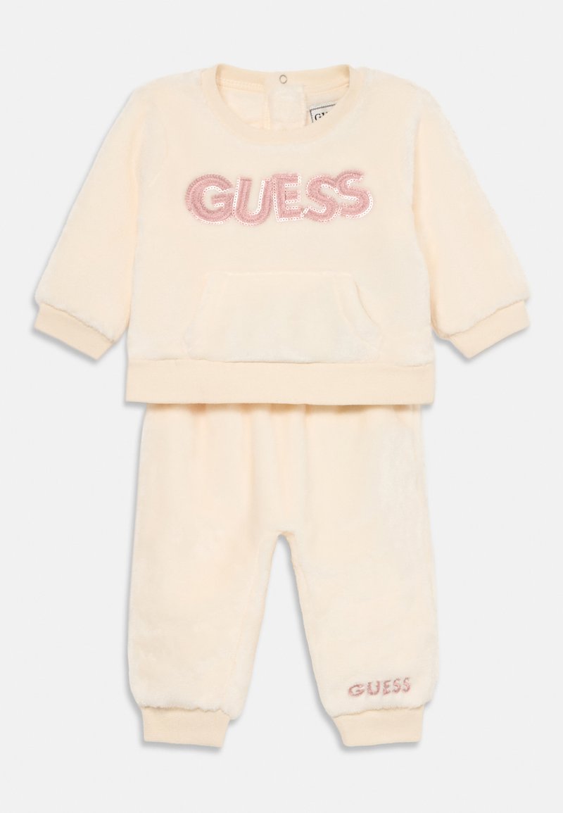 Vauvamallin kreemivärvi plush pitkähihainen collegepaita ja housut, joissa on vaaleanpunainen paljetein koristeltu "GUESS" logo rinnassa ja housunlahkeessa.