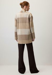 Bēšs un brūns vilnas pleda mētelis ar augstu apkakli un garām piedurknēm, kombinēts ar tumši brūnām plaša piegriezuma biksēm. Gluda tekstūra, vidēja garuma dizains.