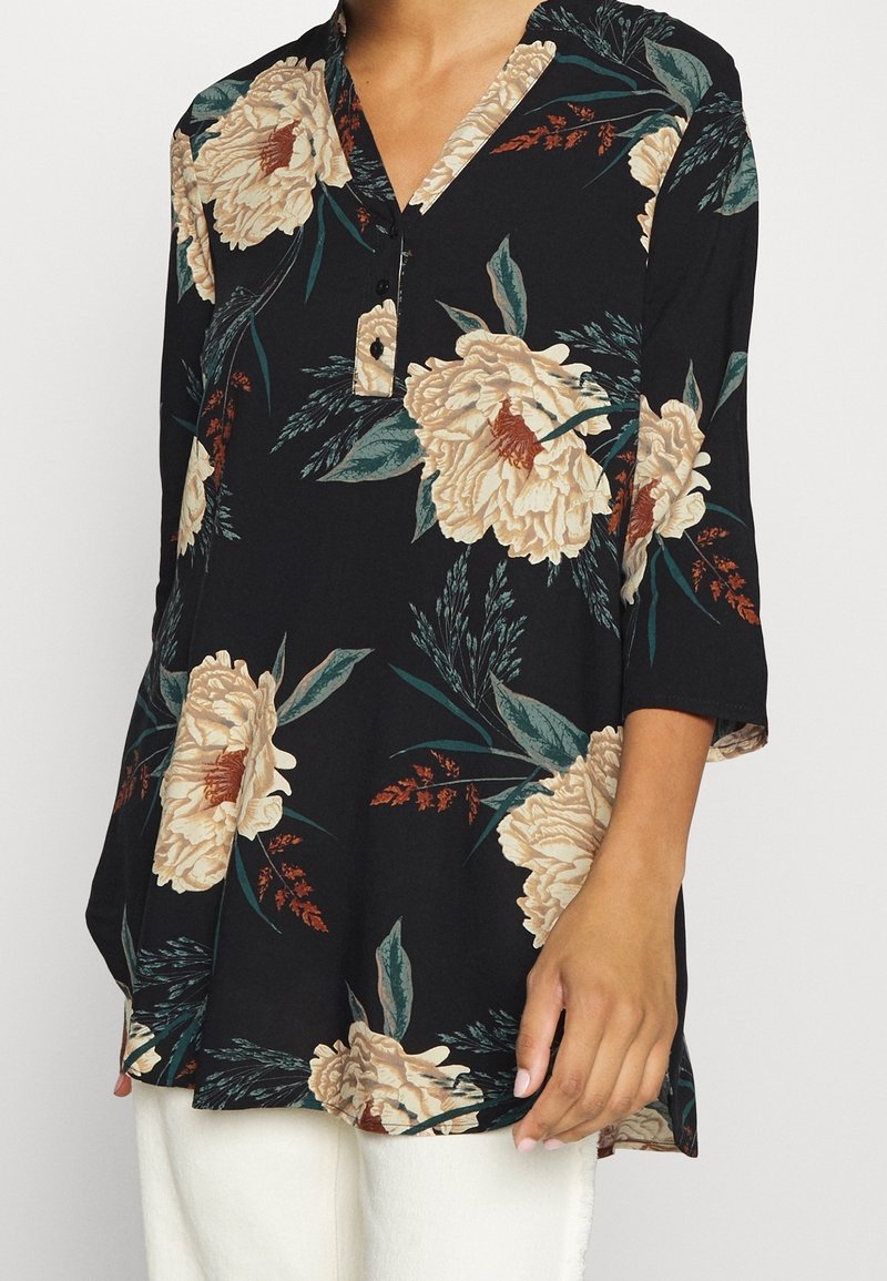 Svart tunika med blommönster som har stora beige blommor och gröna blad. Knäppt halsringning, trekvartslånga ärmar och avslappnad passform.