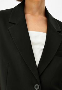 Svart kavaj med slagkrage och enkel knappstängning. Skräddarsydd passform med en texturerad vit topp under. Minimalistisk design.