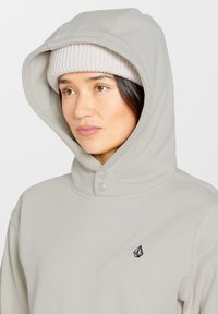 Lichtgrijze hoodie met een grote, zachte capuchon en twee knoopsluitingen bij de kraag. Voorzien van een klein zwart logo op de borst.
