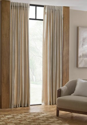 Tende beige chiare pendono da una finestra incorniciata da pannelli in legno, accanto a una poltrona beige con un cuscino in una stanza illuminata delicatamente.