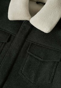 Veste en laine vert foncé avec poches à rabat à l’avant et un col en polaire blanche épais.