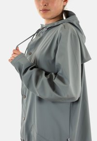 Giacca impermeabile grigio chiaro con cappuccio, bottoni automatici e coulisse regolabile. Presenta una vestibilità ampia e tasche laterali. Tessuto liscio.