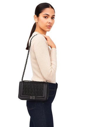Donna con capelli scuri raccolti in una coda di cavallo, che indossa un maglione a coste beige e jeans scuri, mentre porta una borsa a spalla nera trapuntata con tracolla a catena.