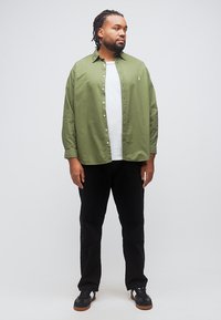 Άνδρας με κοτσίδες κοιτάζει δεξιά, φορώντας πράσινο πουκάμισο με κουμπιά πάνω από λευκό t-shirt, μαύρο παντελόνι και μαύρα sneakers, σε απλό φόντο.