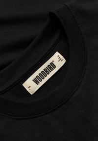 Μαύρο t-shirt με στρογγυλή λαιμόκοψη και μπεζ ετικέτα ρούχου με την επιγραφή "WOODBIRD" και λεπτομέρειες μεγέθους ραμμένες μέσα στη λαιμόκοψη.