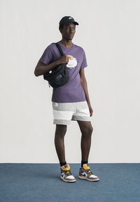 Mand iført en lilla T-shirt med kaffekoptryk, grå og hvide shorts, sort Nike-kasket, sort skuldertaske og Nike-sneakers.