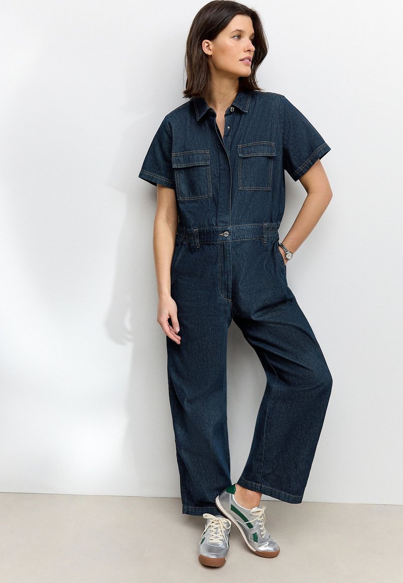 Femme portant une combinaison en denim foncé à manches courtes et poches avant, associée à des baskets argentées et vertes, debout contre un mur blanc.
