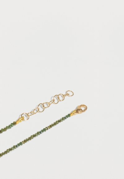 Χρυσός γάντζος και αλυσίδα επέκτασης προσαρτημένα σε πράσινο κολιέ με μικρές διαμαντωμένες πέτρες σε λευκό φόντο.