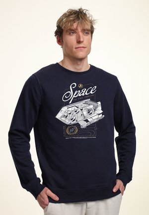 Młody mężczyzna w granatowej bluzie z kapturem z białym statkiem kosmicznym i napisem „Space The Final Frontier”, ręce w kieszeniach, neutralne tło.