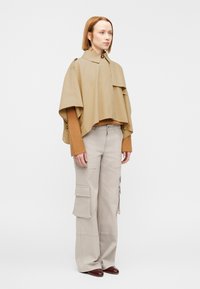 Cape courte couleur fauve avec de larges manches, portée sur un pull marron côtelé. Assorti à un pantalon cargo beige clair avec des poches latérales. Chaussures marron.