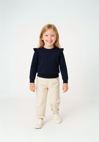 Dunkelblaues Strickpullover mit Rüschendetails an den Schultern, kombiniert mit hochgekrempelten cremefarbenen Hosen und weißen Sneakers.