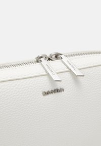 Bílá kožená taštička s texturou, stříbrnými dvojitými jezdci na zipu a logem Calvin Klein ve stříbrném kovu na přední straně.