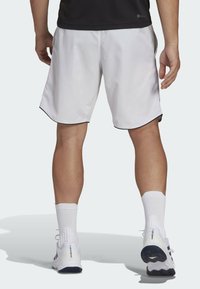 Man som bär vita sportshorts, vita strumpor och vita Adidas GameCourt 2 sneakers, står med ryggen mot kameran.