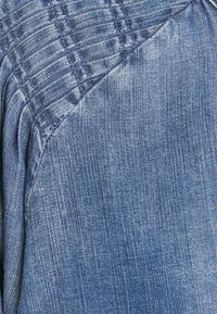 Tecido de ganga em azul claro com uma superfície texturizada. Apresenta smocking integrado, criando um efeito franzido ao longo da costura.