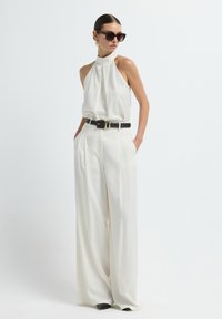 Crème haltertop gecombineerd met hooggeurende, wijdvallende crèmekleurige broek. Zwarte riem met gouden gesp en donkere zonnebril. Glad, lichtgewicht materiaal.