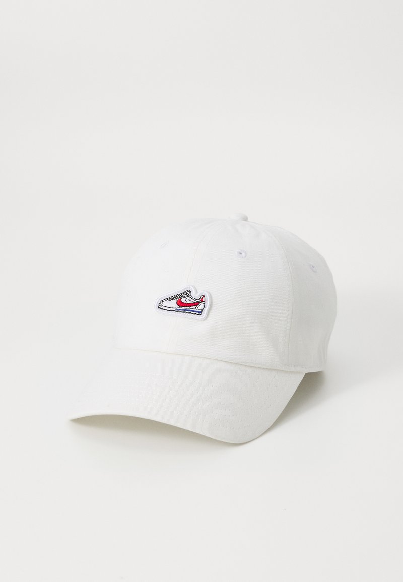Hvit bomullslue med buet skjerm og justerbar bakre stropp. Har et brodert sneakers-logo i rødt, svart og grått på fronten.