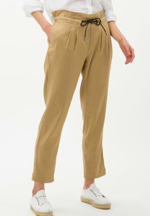 Pantalon beige coupe décontractée avec taille à cordon, porté avec des baskets blanches et une chemise blanche à manches retroussées.