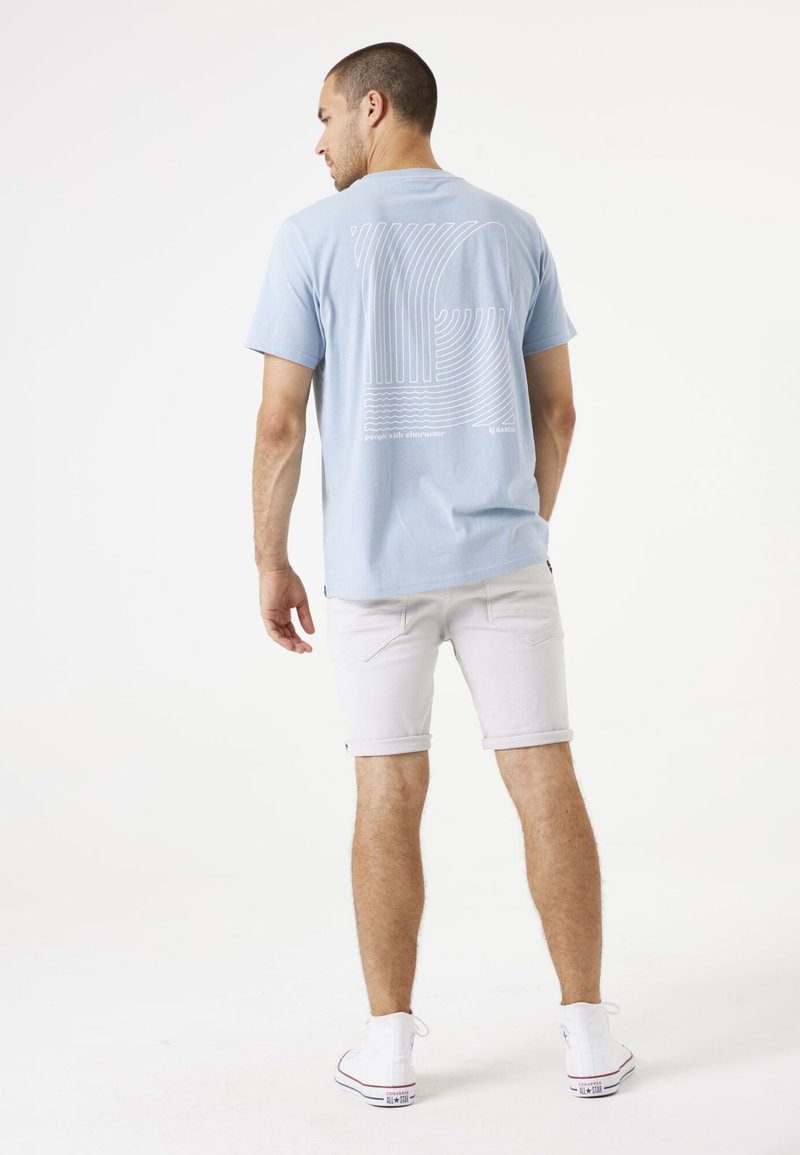 Camiseta azul claro con diseño de arte lineal blanco en la parte posterior, combinada con pantalones cortos blancos enrollados y zapatillas blancas. Diseño simple y casual.