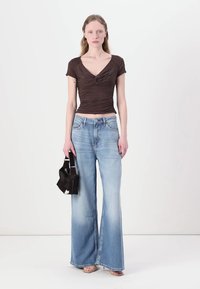 Brown fitted top with front ruching, paired with light blue flared jeans that fade to a lighter tone. Model holding a small black bag.
