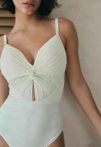 Maillot de bain une pièce vert clair avec tissu plissé, bonnets triangulaires, et détail noué à la taille avec des accents découpés.