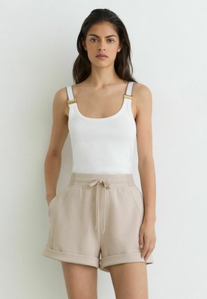 Junge Frau mit langen dunklen Haaren trägt ein weißes Tanktop mit goldenen Schnallträgern und beige Shorts mit Kordelzug, steht an einer weißen Wand.