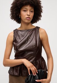 Hnedý, textúrovaný top bez rukávov s peplum lemom. Materiál má lesklý, pokrčený povrch. Skombinovaný s čiernou clutch taškou.