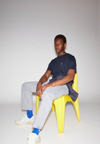 T-shirt marine, jeans gris clair, et baskets blanches avec des accents pastel. Modèle assis sur une chaise en plastique jaune vif avec une chemise rayée nouée à la taille.