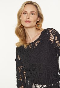 Top din croșetă neagră, cu modele și texturi intricate. Mâneci rotunde, deschise și un decolteu larg îmbunătățesc designul.