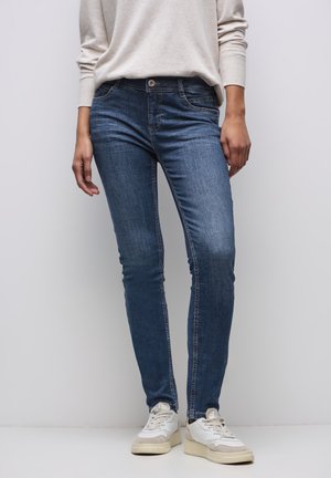 Vaqueros ajustados de mezclilla azul con cintura de altura media. Presentan bolsillos delanteros, cremallera y un sutil desgaste en los muslos. Combinados con zapatillas blancas.