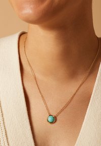 Gouden ketting met een ronde turquoise hanger, gezet in een gestructureerde bezel. Draagt tegen een lichtbeige top, waardoor de kleur wordt benadrukt.