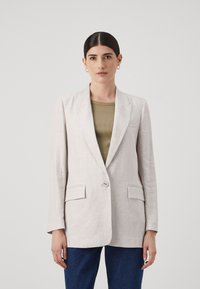 Blazer in lino beige chiaro con rever a punta, chiusura con un solo bottone, due tasche frontali e vestibilità rilassata, abbinato a un top verde oliva.
