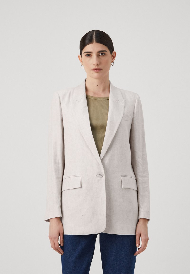 Blazer in lino beige chiaro con rever a punta, chiusura con un solo bottone, due tasche frontali e vestibilità rilassata, abbinato a un top verde oliva.