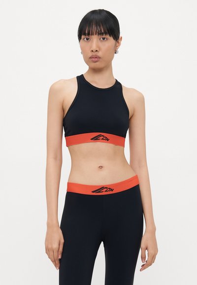 Soutien-gorge de sport noir avec bande orange arborant le logo, associé à des leggings noirs assortis et une ceinture orange. Tissu lisse et ajusté.
