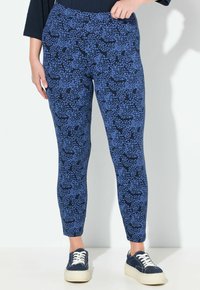 Persona che indossa leggings a pois blu e neri e sneakers platform blu navy, in piedi contro un muro bianco.