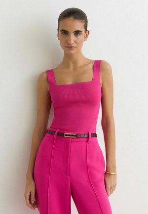 Žena s tmavými vlasy upravenými dozadu, oblečená v jasně růžovém tílku bez rukávů a k němu ladících vysokých kalhotách s černým páskem.