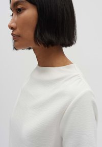 White textured top with a high neckline, featuring soft, subtle patterning. The design includes slight shoulder detailing and a relaxed fit.