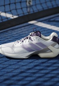 Chaussure de sport Adidas blanche et violette sur un court de tennis bleu à côté d'un filet noir projetant une ombre sur la chaussure et le sol.