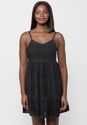 Schwarzes ärmelloses Kleid mit einem figurbetonten Oberteil. Eine Augenlochstickerei schmückt die Taille und den Saum, mit einem Rüschensaum. Glatte Textur.