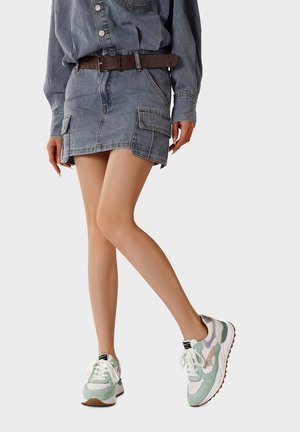 Donna che indossa una gonna cargo in denim azzurro chiaro, camicia in denim abbinata, cintura marrone e sneakers bianche e verdi, in piedi con le gambe incrociate.