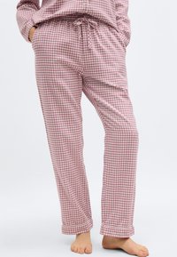 Άτομο που φοράει ροζ και λευκό καρό πυτζάμα παντελόνι με τα χέρια στις τσέπες, στεκόμενο ξυπόλητο μπροστά σε λευκό φόντο.