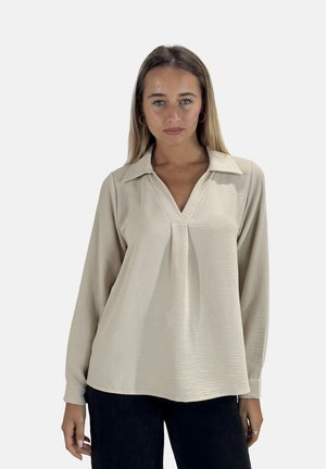 Chemise beige à manches longues avec un col, présentant un décolleté en V et une texture subtile. Assortie à un pantalon sombre.