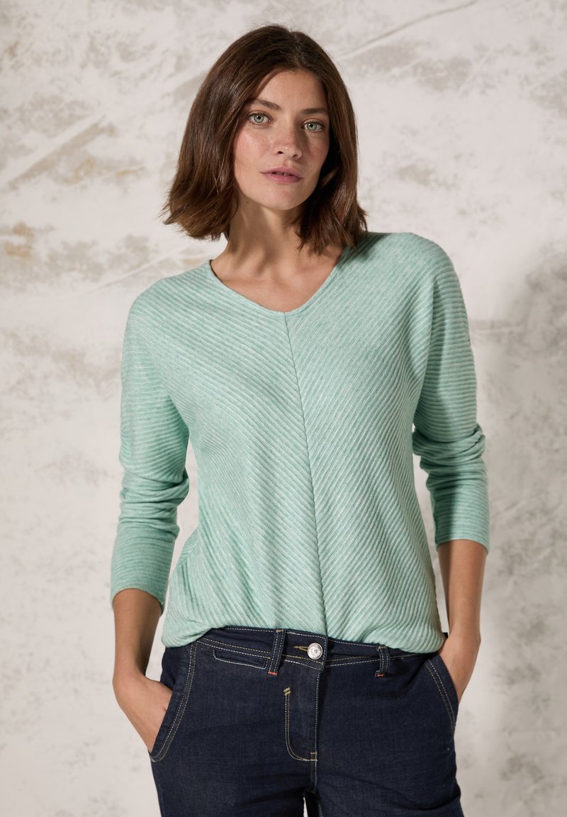 Maglia a strisce verde menta con scollo a V e maniche lunghe, realizzata in tessuto morbido, indossata con jeans scuri dotati di bottoni e tasche.