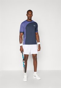 Conjunto atlético masculino composto por uma camisa de manga curta azul-marinho e roxa e calções brancos, segurando uma raquete de ténis. Tecido suave, design moderno.