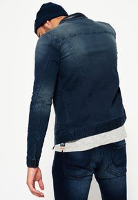 Mörk denimjacka med texturerad yta, figursydd design och kontrasterande sömmar; har en dragkedjeförsedd ficka och etikett med varumärke i nederkant.