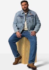 Chaqueta de mezclilla azul claro con parches, camisa crema debajo, jeans azules y botines marrones con cordones, sentado sobre un bloque de madera.