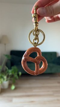 Brown pretzel-shaped keychain made of glossy material, featuring a gold metal clasp and ring for attachment. Interior has small white specks.