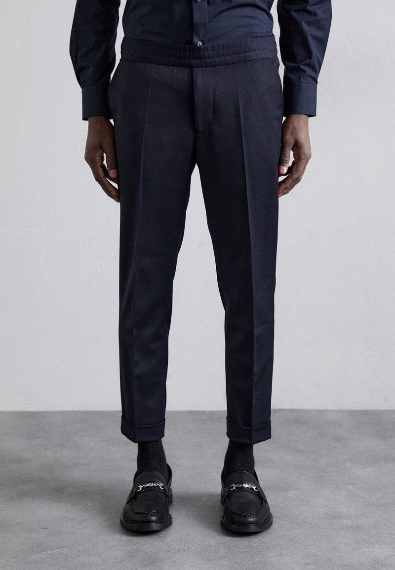 Homme portant un pantalon ajusté bleu marine foncé avec ourlets retroussés, des chaussettes noires, des mocassins noirs avec un détail en chaîne argentée, et une chemise boutonnée bleu marine.